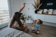 © qunica.com - A mother sits on the floor with her young child, both engaged in a playful moment as the child reaches for a wooden toy. Bright, cozy living space with plants and family photos.