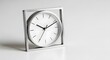 © Raffaza - A minimalist desk clock, a white circular dial with black hands sits within a square silver frame, on a white surface