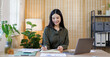 © David - Young asian woman working on laptop in modern office interior with shelves plants and stationary creating a cozy yet professional workplace atmosphere indoors.