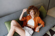 © SHOTPRIME STUDIO - Cheerful young woman with curly hair relaxing on a sofa in a cozy indoor setting, wearing an orange shirt and white shorts, embodying a sense of tranquility and comfort