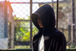 © Aoy_Charin - Mysterious teenage boy in a black hoodie standing in front of a chain-link fence. Urban youth, crime, and anonymity concept.