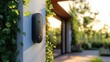 © Tatyana - modern black video doorbell mounted on white wall surrounded by green leaves with a blurred garden and house exterior in the background during golden hour