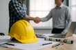 © diproduction - Yellow hard hat on architectural blueprints with two professionals shaking hands in office, symbolizing construction business agreement concept. Ai generative