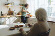 © shurkin_son - Rare view of senior lady sitting at kitchen table, having herbal tea looking at nurse or caregiver cutting pumpkin standing next to counter, having nice conversation while social worker cooking