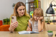 © Andrii Iemelianenko - Mother and daughter at home watch online lesson on smartphone write notes in notebook follow tasks. Woman and girl at table check exercise mark answer fix mistakes learn rule practice focus pace well