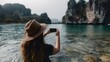 © Bussakon - A woman wearing a straw hat takes a photo with her smartphone capturing the serene natural beauty of tropical islands and clear blue waters