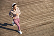 © DragonImages - Young adult Caucasian woman jogging outdoors on wooden boardwalk wearing headphones, looking ahead with focused expression, engaging in fitness activity, exercising alone
