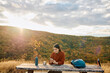 © we.bond.creations - Woman taking a break while hiking, sitting at a picnic table and using phone during sunset in the mountains