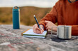 © we.bond.creations - Person's hand writing in a spiral notebook, journaling thoughts on a rustic wooden table with a traveling mug and water bottle