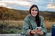© we.bond.creations - Young woman smiling, holding smartphone, relaxing outdoors during autumn hiking trip, enjoying connection and peace