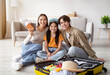 © Prostock-studio - A cheerful family sits together on the floor in a living room, surrounded by a suitcase filled with travel items. They are excited for their upcoming trip.