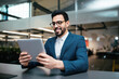 © Prostock-studio - A man in a blazer sits at a sleek desk in a contemporary office. He smiles as he interacts with his tablet, surrounded by glass walls and plants, showing a productive environment.