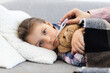 © sofiko14 - A young girl lies on a couch with a thermometer in her mouth, being cared for by a parent. She is wrapped in a blanket with a teddy bear.