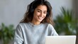 © Yuliia - Young Hispanic woman in gray sweater smiling while working on laptop at home office, with green plants in background. Natural lifestyle portrait for remote work and productivity.