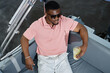 © TrueFrame Collective - Black young adult man relaxing on boat holding cold drink with lemon slices, wearing sunglasses, sitting on cushioned seat, looking away from camera, enjoying leisure time outdoors