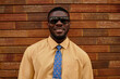 © TrueFrame Collective - Portrait of young adult Black man smiling and wearing sunglasses while standing against brick wall, facial hair visible, looking directly at camera, formal attire evident