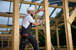© Василь Івасюк - Construction worker measuring wooden beam with tape measure at building frame. Builder in safety helmet working on wooden house construction site outdoors.