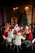 © deagreez - Family Christmas dinner around a festive table with Santa hats in a cozy loft with a tall Christmas tree and warm lights