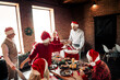 © deagreez - Friends and family gather for a cozy Christmas dinner indoors wearing Santa hats sharing laughs and food around a festive table