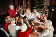 © deagreez - Family gathers around a festive Christmas dinner to toast with champagne by a decorated tree and warm lights creating a cozy home holiday atmosphere