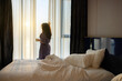 © shine - Morning of Beautiful asian middle aged woman opening curtains in bedroom  enjoying view of sunrise, strong color clouds light rays and other atmospheric effect from window while having a cup of coffee