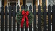 © Lasvu - A black wooden fence with wood palings and an evergreen wreath decorated with a red ribbon adorns the front entrance of a historic yellow and white house with a black door during Christmas.