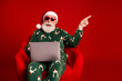 © deagreez - Santa in festive pajama sits in a red chair with a laptop wearing sunglasses and points to celebrate Christmas Eve from a warm home setting