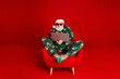 © deagreez - Santa in green pajama sits in a red chair using a laptop for Christmas online shopping and holiday fun in a bright red studio with merry festive vibes
