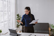 © amnaj - Asian businesswoman presenting during a virtual meeting, standing and holding documents while using a tablet at her home office