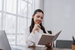 © amnaj - Asian woman smiling, holding a notebook, and writing notes while working on business planning in a modern office