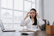 © amnaj - Young Asian woman experiencing severe headache and high stress while sitting at her office desk, looking tired and overworked