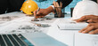© InfiniteFlow - A focused team of engineers collaborates on detailed construction plans in an office environment, using safety helmets and modern technology for effective project execution. SACTR