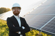 © Serhii - Smiling Engineer wearing white hardhat posing with arms crossed in front of solar panels