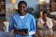 © DragonImages - Portrait of young adult Black woman wearing medical scrubs smiling at camera holding clipboard and pen, senior Black woman sitting in background using walking cane in living room, during patient visit