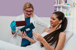 © Dusko - Cosmetologist in blue gloves and lab coat presenting various skincare products to a smiling female client lying on a treatment bed in a modern beauty clinic