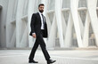 © Pete - Bearded man in elegant black suit strides confidently past modern building. Pro male walks with purpose, exuding success, determination. Business executive on city street, focused on career goals.