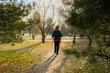 © tiena - A full-length portrait of a Caucasian woman wearing a jacket and hat with Nordic walking poles walks through a pine forest in a city park, back view