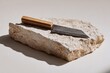 © Spin - A rustic, dark-metal chef's knife with a light wood handle rests on a rough, light-beige stone.  Subtle shadows enhance the minimalist aesthetic