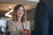 © DIMENSIONS - Smiling businesswoman in a beige blazer engaging in a confident handshake in a modern office setting representing professional networking, partnership, and career success