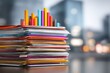 © SY - Stack of colorful paper folders with graph on top, in blurred cityscape. It represents data analysis, paperwork, and organization for business use.