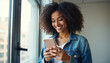 © Maryna - Happy black woman with curly hair smiles using smartphone near window. Female person texting on cellphone indoors. Young adult female with mobile device at home.
