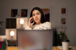 © Studio Marmellata - Young Arab female remote employee in casual clothes speaking via smartphone with client while working on project remotely using laptop