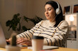 © Studio Marmellata - Young concentrated female freelancer in headphones sitting at table using computer while working remotely at home in modern apartment