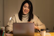 © Studio Marmellata - Positive young Arab woman in casual sweater sitting at table with laptop and making a video call on project during evening time