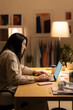 © Studio Marmellata - Positive young Arab woman in casual sweater sitting at table with laptop and working online on project from home during evening time