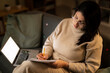 © Studio Marmellata - Concentrated Arab female in casual beige outfit and eyeglasses sitting on sofa with documents using laptop while working online at home