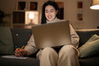 © Studio Marmellata - Arab female freelancer in casual wear and glasses typing information on laptop while working remotely sitting on sofa at home in evening