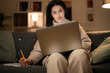 © Studio Marmellata - Arab female freelancer in casual wear and glasses typing information on laptop while working remotely sitting on sofa at home in evening