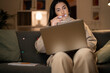 © Studio Marmellata - Arab female freelancer in casual wear and glasses typing information on laptop while working remotely sitting on sofa at home in evening