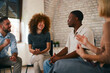 © djile - Smiling young woman with curly hair looks at an African American man speaking expressively during a positive and supportive multi-ethnic group therapy session indoors.